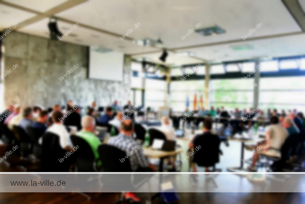 Eine Gruppe von Personen, die in einem modernen Konferenzraum mit großen Fenstern sitzen und an einer formellen Sitzung oder Diskussion teilnehmen.