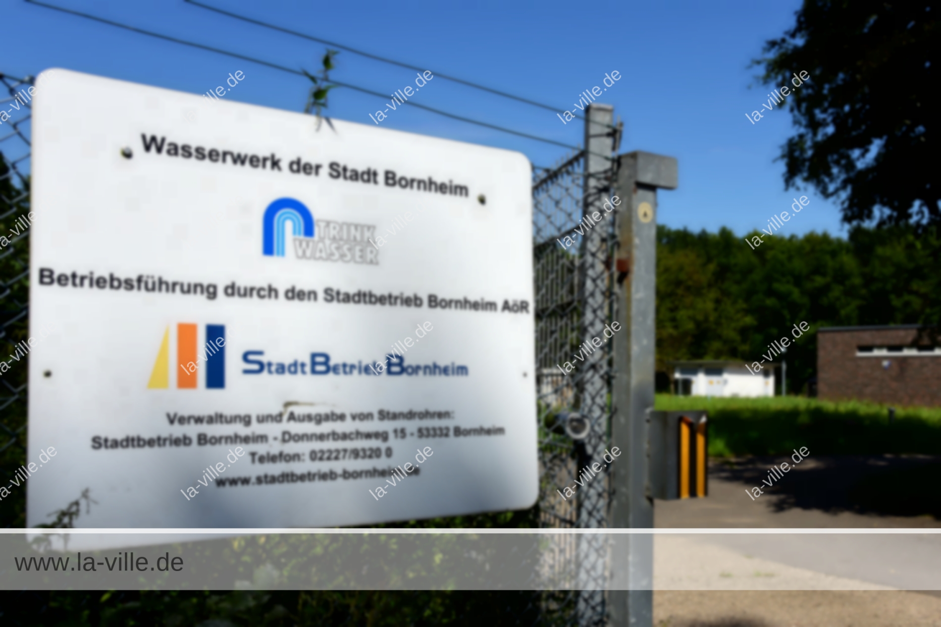 Schild am Eingang des Wasserwerks Bornheim mit Angaben zur Verwaltung und zu den Ansprechpartnern in deutscher Sprache, im Hintergrund ein Gebäude und eine Grünanlage.